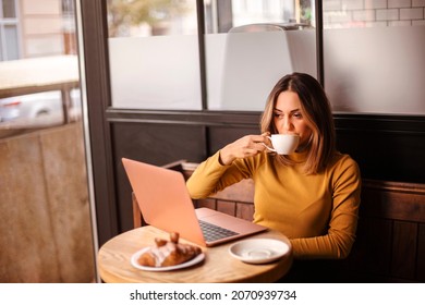 Young woman freelancer drinking coffee and eating croissant while working remotely on laptop computer in cafe, caucasian female enjoying breakfast during online remote work in coffeeshop
