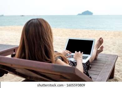 Young Woman Freelancer Sitting At The Table On Ocean Background, Using Laptop With Black Screen On The Beach. Girl Freelancer Working. Work Outside Office Concept.