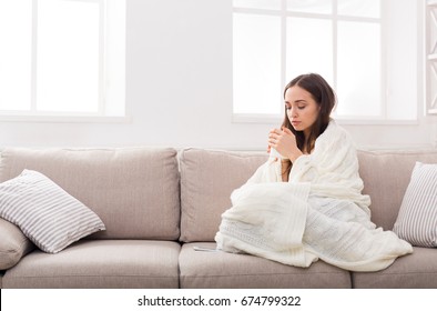 Young Woman Freezing At Home. Warm Up Wrapped In Blanket And Drinking Coffee With Mobile Lying Next To Her, Copy Space