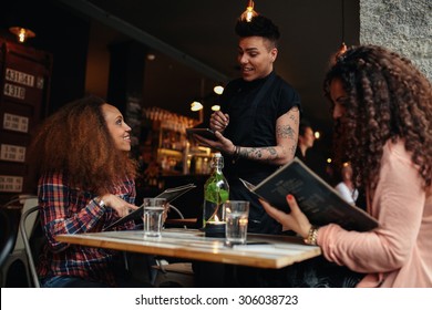 Young woman with a friend ordering to waiter holding digital tablet. Two women sitting at cafe holding menu card giving an order to male waiter.