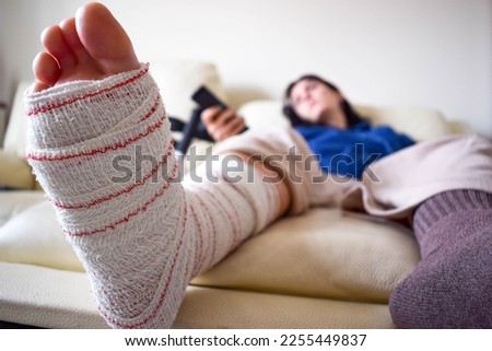 Young woman frustrated on couch at home with crutches and orthopedic plaster watching TV. Fracture of the broken leg foot or knee. Concept of rehabilitation and healing. Orthopedics and Traumatology.