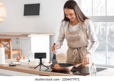 Young Woman Frying Pancakes While Following Video Tutorial In Kitchen