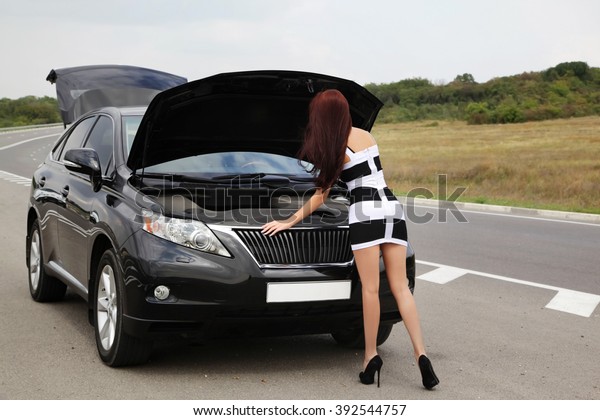 Young\
woman in full length standing on a road near broken car. Looking at\
the hood of the car trying to identify the\
problem