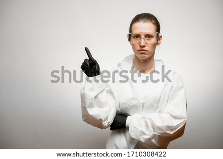 young woman in full medical protective clothing and goggles on her face stands on white background and shows hand to side