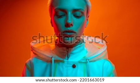 Young woman in futuristic outfit with short hair standing under turquoise neon light against orange background