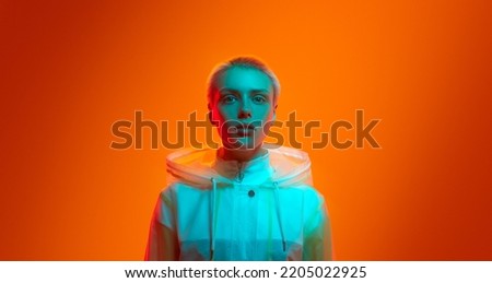 Young woman in futuristic raincoat with shot hair looking at camera while standing under turquoise neon light against orange background