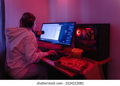 Young Woman Gamer Or Software Developer Sitting At The Modern Personal Computer And Eating Junk Food At Night In Room With Neon Lights