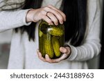 A young woman, a gardener, a cook, holds a glass jar with canned cucumbers, preserves, in her hands in the kitchen, opening the lid. Food photography.