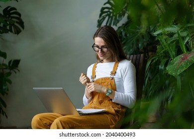 Young Woman Gardener Or Student Watch Online Webinar On Home Gardening And Florist Small Business Plan And Houseplants Care On Laptop Studying Make Note For Work On Greenhouse And Development Strategy