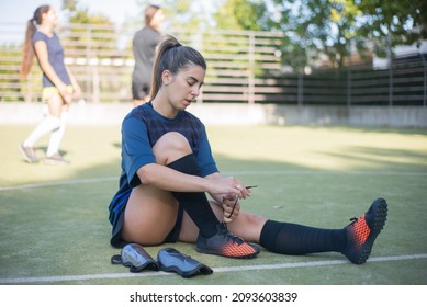 Young Woman Getting Dressed For Training. Female Player Sitting On Grass, Tying Laces Of Black Football Boots, Concentrated. Football, Sport, Leisure Activities Concept