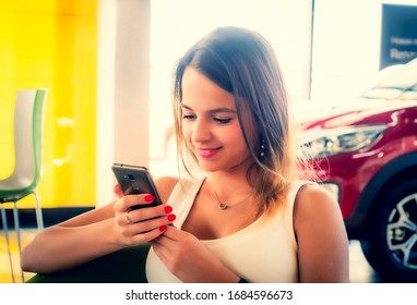 Young Woman Getting Happy News In Her Phone In The Car Dealership. Happy Face Of A Girl Sitting On A Visitor Chair With Her Phone In Hands.