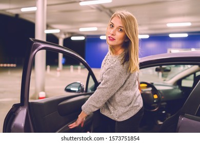 Young Woman Getting Out Of The Parked Car Looking To The Camera Smiling On The Parking In The City At Night