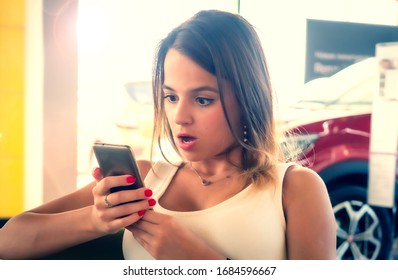 Young Woman Getting Shocked By News In Her Phone In The Car Dealership. Shocked Face Of A Girl Sitting On A Visitor Chair With Her Phone In Hands.