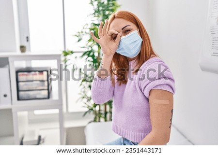 Young woman getting vaccine showing arm with band aid smiling happy doing ok sign with hand on eye looking through fingers 