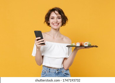 Young Woman Girl In Casual Clothes Hold In Hand Makizushi Sushi Roll Served On Black Plate Japanese Food Using Mobile Cell Phone Isolated On Yellow Background Studio Portrait People Lifestyle Concept