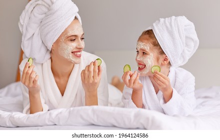 Young woman and girl with cucumbers smiling and looking at each other while relaxing on bed during spa procedure at home