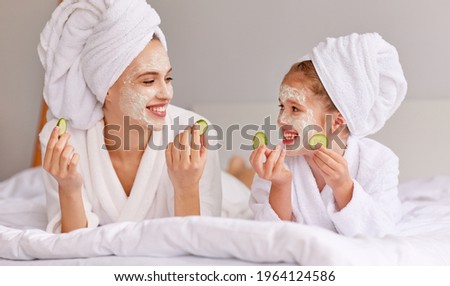 Young woman and girl with cucumbers smiling and looking at each other while relaxing on bed during spa procedure at home
