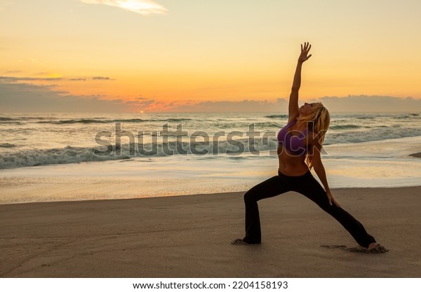 Young woman girl female in a warrior\
position practicing yoga on a beach at sunrise or\
sunset