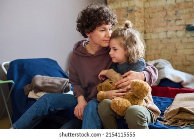 Young woman giving hug to her cute little son with brown soft teddybear while both sitting on sleeping place prepared for refugees