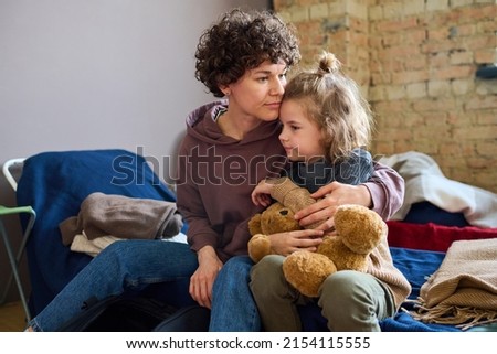 Young woman giving hug to her cute little son with brown soft teddybear while both sitting on sleeping place prepared for refugees