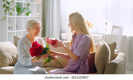 Young Woman Giving A Red Flowers To His Elderly Mother In A Home Interior. Smiling Girl With Her Mother. Happy Birthday