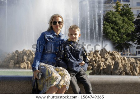 young woman in glasses and a boy sit on the background of the city fountain