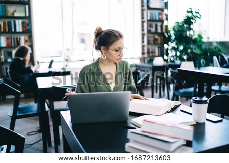 Young woman in glasses and elegant clothes sitting at desk among printed literature and laptop while  reading book in modern library