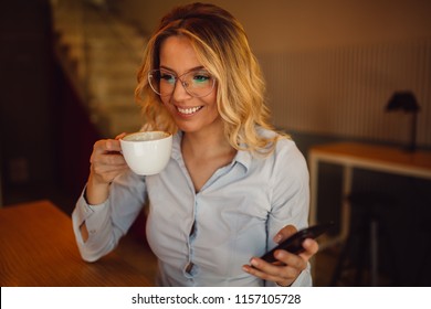 Young woman with glasses is holding smartphone in hand while drinking coffee in cafe at coffee break. - Shutterstock ID 1157105728