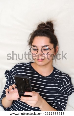 A young woman in glasses lies on the bed and looks at the phone communicates to study at work