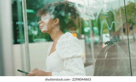 Young woman in glasses looking at a smartphone while standing at a bus stop. Positive woman using mobile phone outdoors in urban background. - Powered by Shutterstock