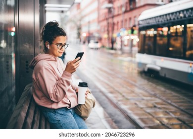 Young woman with glasses and mobile phone waits for bus and gets into salon on public transport stop against large city blurry street in rainy evening side view