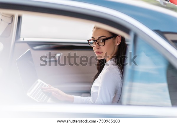 Young woman in glasses sitting in a\
backseat of a car with laptop and looking at\
camera