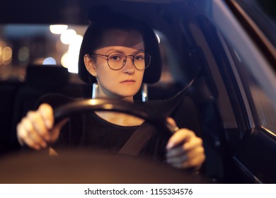 Young Woman With Glasses At The Wheel Of The Car. Night City And Light Of Street Lamps. Front View. Serious, Focused Look. He Drives A Car On The Road, Overtakes.