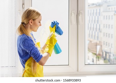 Young woman in gloves and apron washing window pane at home with rag and spray detergent. Blue yellow color. Cleaning concept.