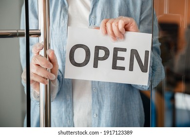 Young Woman In Gloves Hangs On Front Door Of Cafe Reopen Sign. Sign Open Welcome On Shop Entrance Door As New Normal. End Lockdown Coronavirus Covid 19 For Local Business.