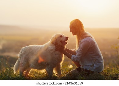 Young woman and golden retriever dog sit on green grass at sunset. Pets friendly travel concept, solar bright light.