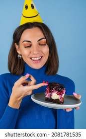 Young Woman In Golf On A Blue Background Celebrates A Birthday, Holds A Piece Of Cake, Happy, Licks Her Finger With Pleasure