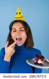 Young Woman In Golf On A Blue Background Celebrates A Birthday, Holds A Piece Of Cake, Happy, Licks Her Finger With Pleasure