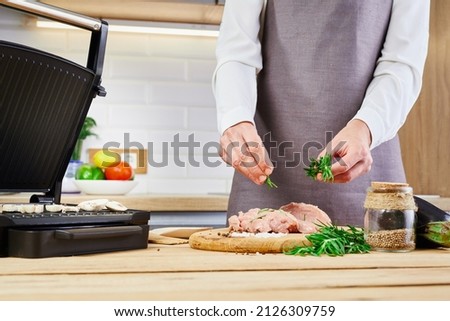 A young woman in a gray apron cooks or decorates raw meat with rosemary in the kitchen.A conceptual woman cooks food at home.