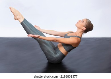 Young Woman In Gray, Two-piece Yoga Outfit, Seated On A Black Floor In A Boat Yoga Pose Against A White Background.