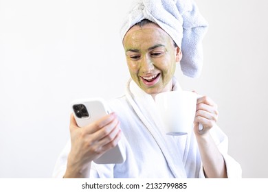 Young Woman With A Green Beauty Mask On Her Face With A Cup And A Smartphone.