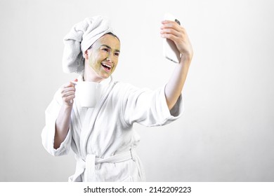 Young Woman With A Green Beauty Mask On Her Face With A Cup And A Smartphone.