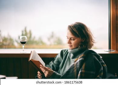 Young Woman In Green Sweater Reads A Book And Drinking Wine Near Window In Her Country House With A View Of The Mountains 