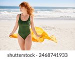 Young woman in green swimsuit holding yellow scarf while walking at beach with copy space. Happy active and healthy woman enjoying summer vacation at sea. Girl with red hair walking at seashore.