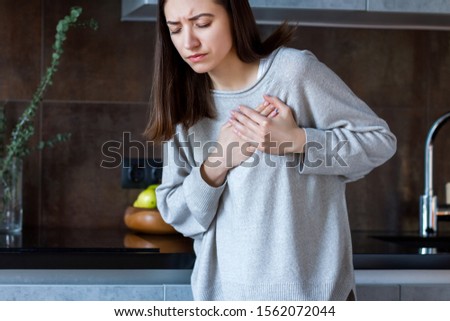 Young woman in grey clothes is holding hands on breast. Brunette girl is feeling bad. Sudden heart attack, myocardial infarction at home. Effect of stress and unhealthy lifestyle concept.