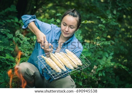 young woman is grilling sweet corn on fire in the forest