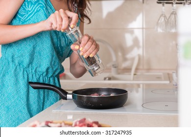 Young Woman Grinding Pepper Onto Cooking Meat In A Frying Pan On The Stove Using A Large Pepper Mill, Close Up Of Her Hands