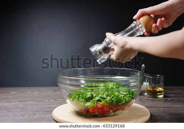 Young woman grinding pepper to salad. Full bowl of\
fresh green salad, tomatoes, cucumber close-up on wood table\
against dark background, rustic kitchen. Salt, olive, sunflower\
oil, pepper on table.
