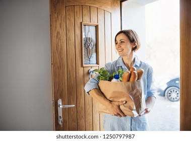 A Young Woman With Groceries In Paper Shopping Bag Walking In Through Front Door.