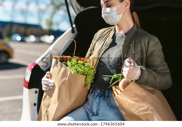 A young woman with grocery bags from a\
supermarket. Car trunk at background. Social distancing: face mask,\
disposable gloves to prevent infection. Food shopping during\
coronavirus Covid 19 epidemic